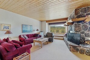 A living room with a stone fireplace, wooden ceiling, and a large window. - Slopeside ski-in/ski-out condo with private hot tub & panoramic views (Big Sky)