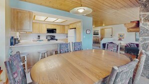 A wooden dining table surrounded by chairs, a kitchen area with wooden cabinets, and a stone accent wall. - Slopeside ski-in/ski-out condo with private hot tub & panoramic views (Big Sky)