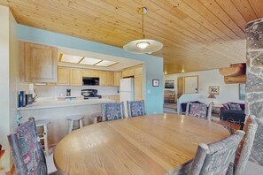 A wooden dining table surrounded by chairs, a kitchen area with wooden cabinets, and a stone accent wall. - Slopeside ski-in/ski-out condo with private hot tub & panoramic views (Big Sky)