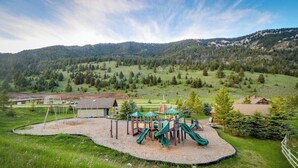 Un parque infantil con tobogán y columpios, rodeado de grava, con colinas verdes y un cielo despejado de fondo.