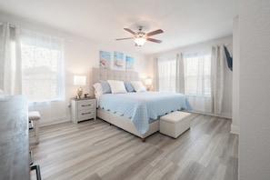Ein Schlafzimmer mit Bett, Nachttischen, Deckenventilator und einem Fenster mit Vorhängen.