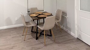 Spiseområde med et bord dækket til fire personer, omgivet af fire stole, med et vindue og en dør i baggrunden.