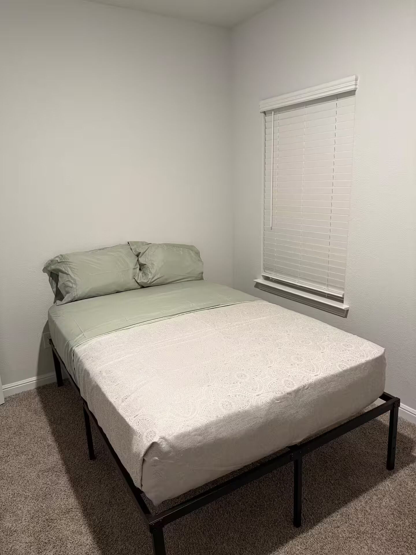 Ein Einzelbett mit weißer Matratze und grünen Kissen in einem Zimmer mit weißen Wänden und einem Fenster mit Jalousien.