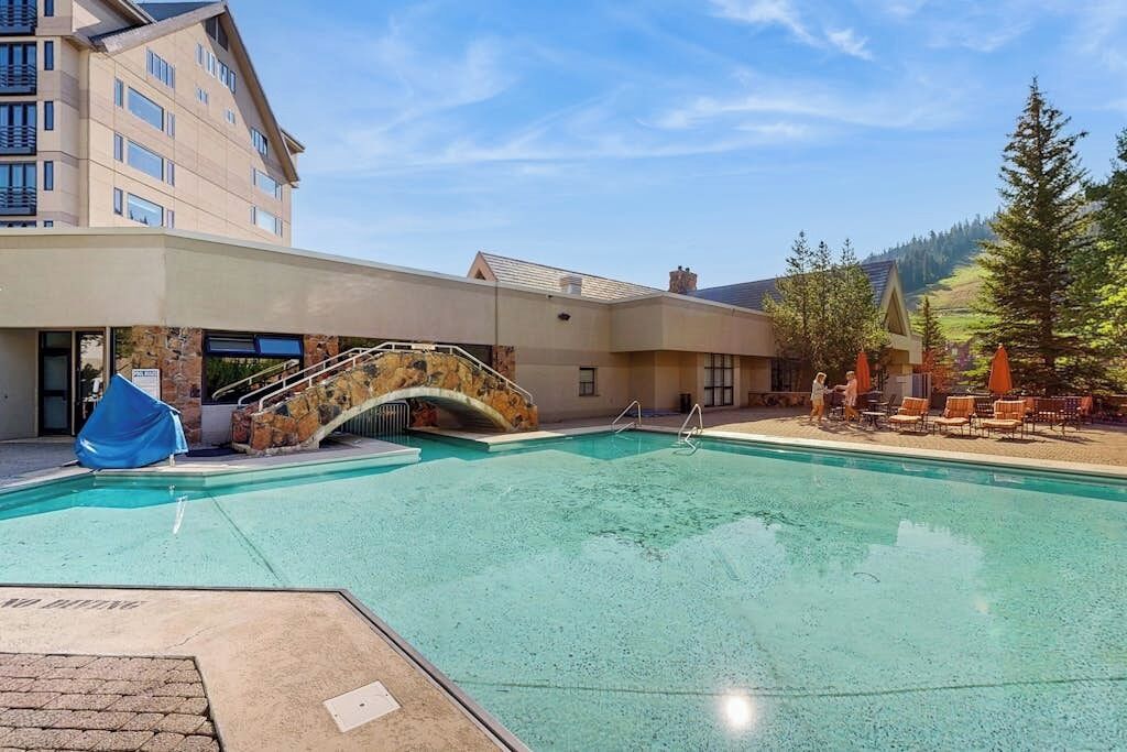 A swimming pool with a stone bridge, outdoor seating, and a building in the background.