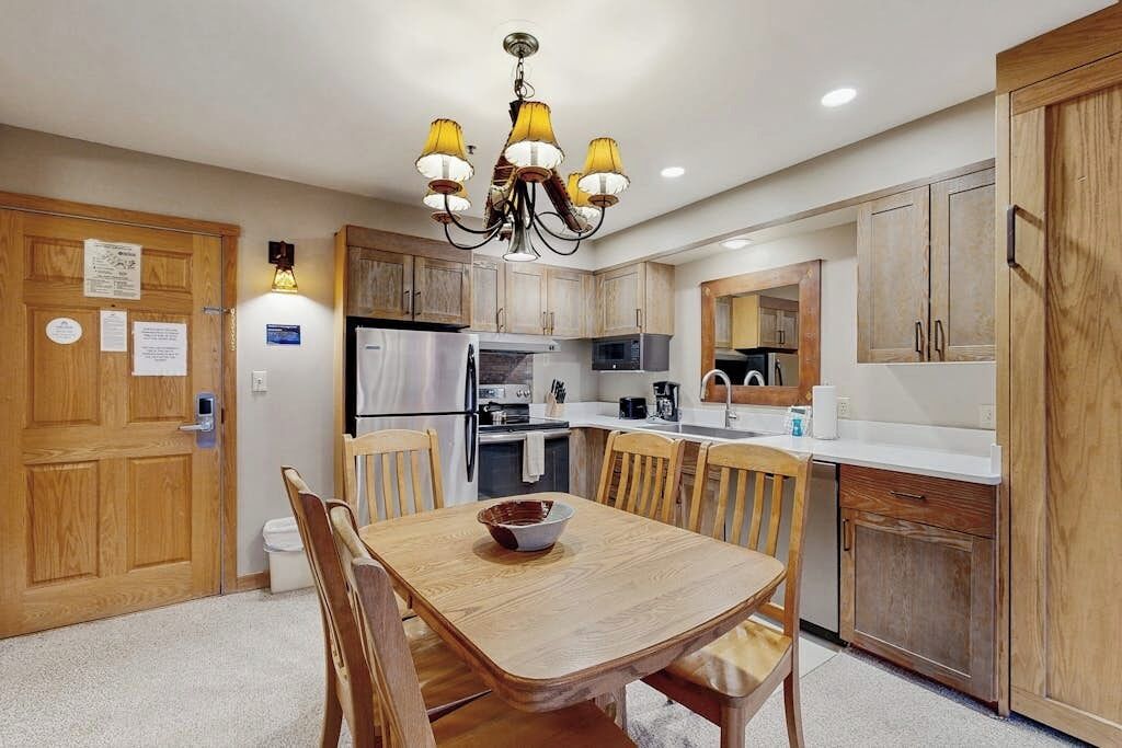 A compact kitchen with wooden cabinets, a dining table with chairs, a chandelier, and a refrigerator.