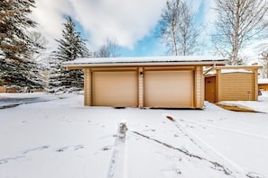 A two-car garage with a snow-covered roof, surrounded by trees and a clear sky.