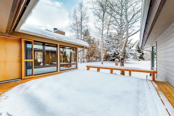 A snow-covered deck with a wooden bench, a glass door leading to a room, and a snow-covered tree in the background.