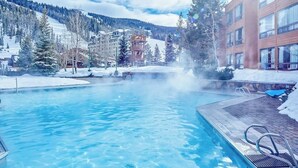 A pool with clear water, surrounded by buildings and snow-covered trees. - Ski-in/out, mountain-view condo with hot tub, pool & gym (Big Sky)