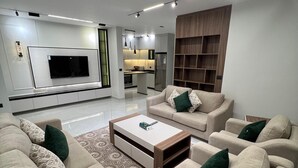 Una sala de estar moderna con un televisor de pantalla plana, sofás color beige, una mesa de centro y una cocina al fondo.