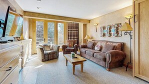A living room with a sofa, armchair, coffee table, and a television on a wooden cabinet. - Ski-in/ski-out ski chalet w/shared hot tub, pool, balcony, WiFi, central A/C (Big Sky)