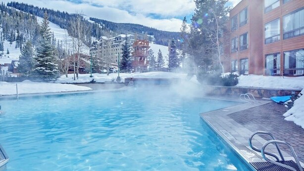 Une piscine à l’eau claire, entourée de bâtiments et de collines enneigées.