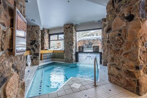 An indoor swimming pool with stone walls, a tiled floor, and a view of the outside. - Ski-in/ski-out condo in the village w/ shared pool/hot tub, sauna, gym & more (Big Sky)