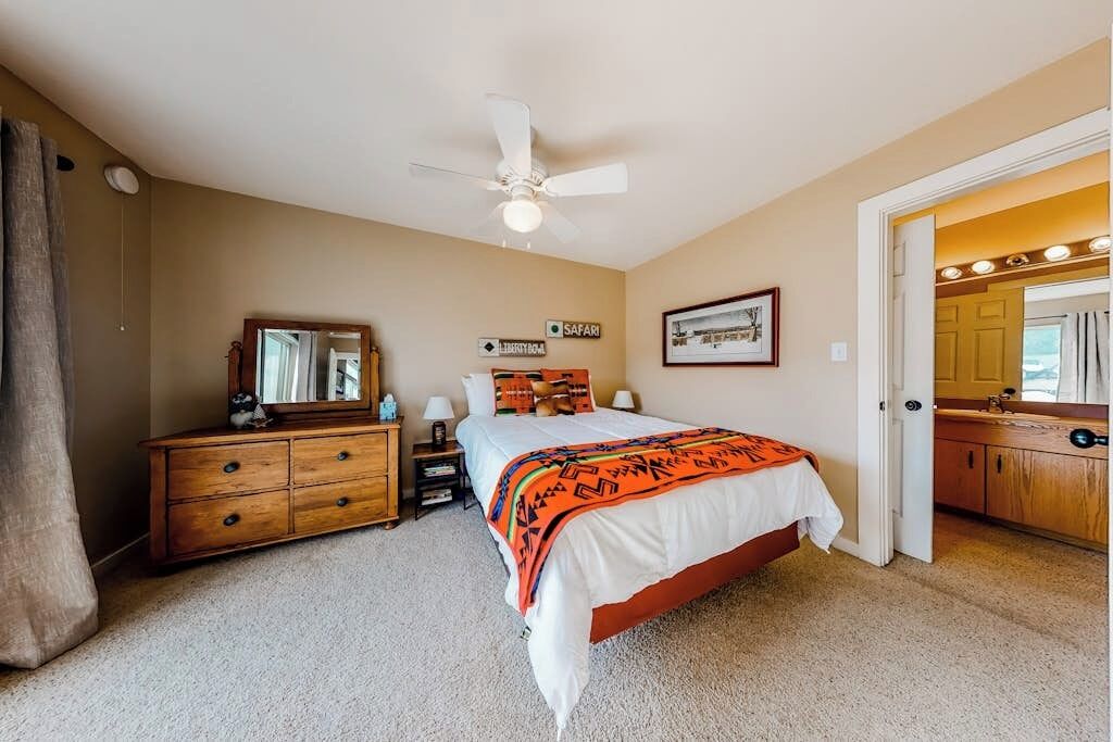 Une chambre à coucher comprenant un lit, une commode, un miroir, un ventilateur de plafond et une porte donnant sur une salle de bain.