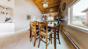 Un espace repas avec des chaises et une table en bois, un plafond avec des poutres apparentes et une fenêtre donnant sur les bâtiments environnants.