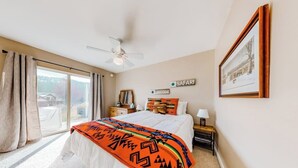 Une chambre à coucher comprenant un lit, un ventilateur de plafond, une table de chevet, une lampe, un tableau encadré et une fenêtre avec des rideaux.