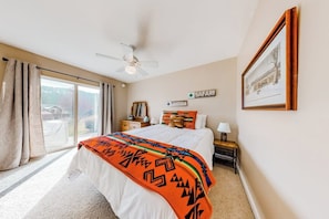 Ein Schlafzimmer mit Bett, Deckenventilator, Nachttisch, Lampe, gerahmtem Bild und Fenster mit Vorhängen.