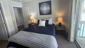 A hotel room with a bed, two bedside tables, a window with curtains, a door, and a wall-mounted artwork. - Immaculate Condo Near Meadow Village | New Carpet, Mountain Views & Dog-Friendly (Big Sky)