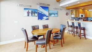 A dining area with a round wooden table and chairs, a kitchen with wooden cabinets, and wall-mounted pictures of mountains. - Serene mountain-view condo with skiing, washer/dryer, gas grill, & patio (Big Sky)