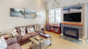 A living room with a sofa, coffee table, fireplace, television, and artwork on the wall. - Serene mountain-view condo with skiing, washer/dryer, gas grill, & patio (Big Sky)