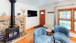 A cozy living room with a stone fireplace, a television, a wooden door, and two blue armchairs. - Modern duplex with gas fireplace, free WiFi, & walkable downtown location (Bozeman)