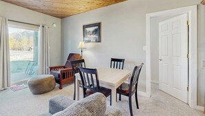 Une pièce avec un plafond en bois, une table à manger avec des chaises, une lampe, un tableau encadré au mur et une porte.