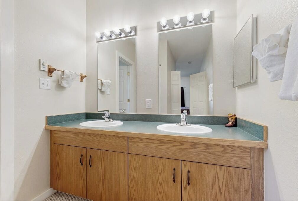 A bathroom with two sinks, wooden cabinets, a mirror with lights, and a towel rack.