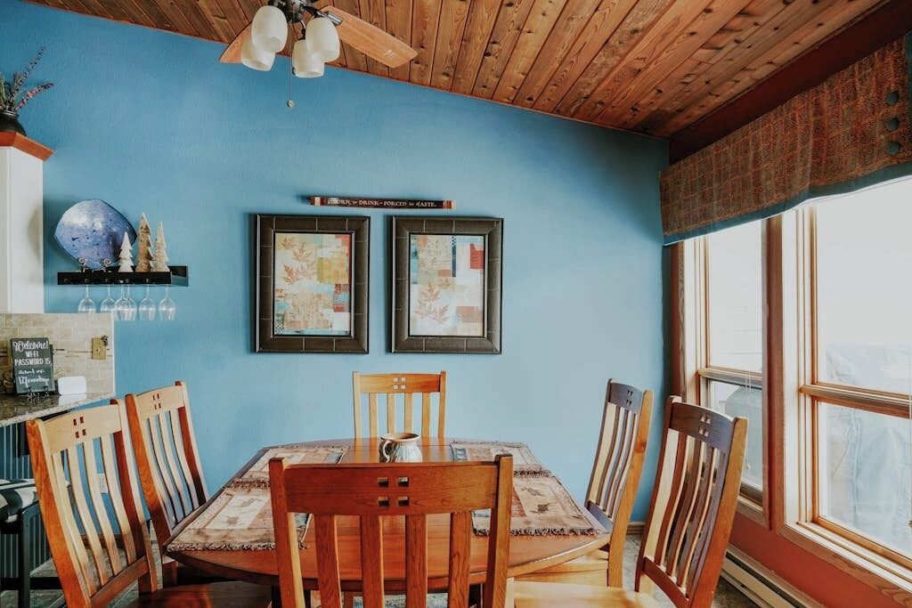 A dining area with a wooden table and chairs, framed pictures on the wall, and a window with curtains.