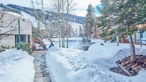 Ein verschneiter Pfad führt zu einem Gebäude mit einer Stein-Fassade, umgeben von Schnee, Bäumen und einem Bergpanorama.