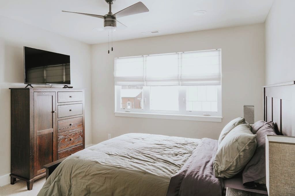 A bedroom with a bed, a dresser, a television, a window, and a ceiling fan.