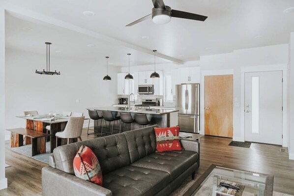 A modern living room with a sofa, dining area, kitchen, and a ceiling fan.