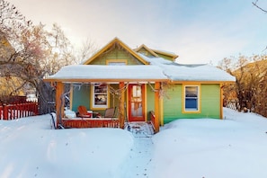 A two-story house with a green exterior, a porch with a red door, and a snow-covered roof. - Charming downtown retreat with stained glass, enclosed yard, and washer/dryer (Bozeman)