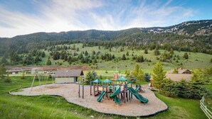 A playground with a slide and swings, surrounded by a gravel area, set against a backdrop of green hills and a clear sky. - Ski-in/out condo near Lake Levinsky with balcony, ski & mountain views (Big Sky)
