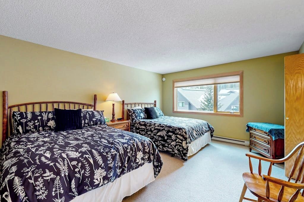 A bedroom with two beds, a nightstand, a chair, and a window with a view of houses.