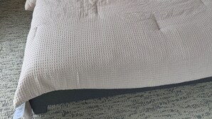 Una cama bien hecha con edredón blanco y almohadas, dos lámparas de noche, una mesita de noche con un libro y una ventana con persianas.