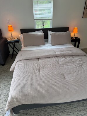 Een net opgemaakt bed met een witte dekbedovertrek en kussens, twee nachtlampjes, een nachtkastje met een boek, en een raam met jaloezieën.