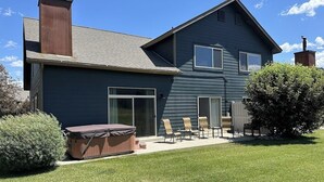 A two-story house with a dark blue exterior, a chimney, a patio with chairs, and a hot tub. - Modern Mountain Getaway with New Hot Tub – Big Sky, Montana (Big Sky)
