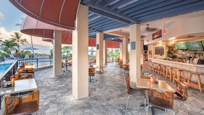 An outdoor bar area with stone flooring, wooden tables and chairs, and a pool in the background.