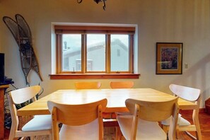 Un coin repas avec une table et des chaises en bois, une fenêtre donnant sur des maisons et un tableau encadré au mur.