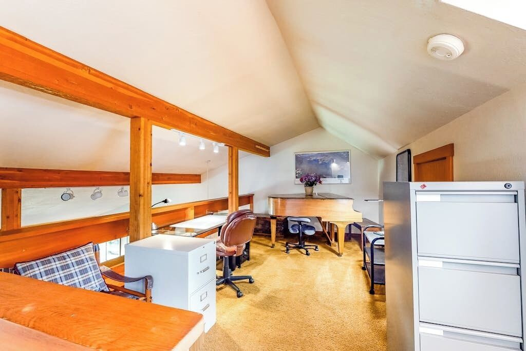 A cozy, well-lit room with a wooden ceiling, a desk with office chairs, a large white refrigerator, and a bed with a plaid pillow.