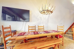 Ein Essbereich mit Holztisch, Stühlen, einer Tischläufer mit Muster, einem Wandfernseher und einem besonderen Kronleuchter.
