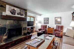 Ein Wohnzimmer mit Kamin, Fernseher, gemusterten Sesseln, einem Couchtisch und einem Bücherregal.