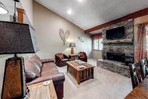 A living room with a stone fireplace, leather sofa, wooden coffee table, and a flat-screen TV mounted on the wall. - Beautiful condo with full kitchen, mountain views (Wilson)
