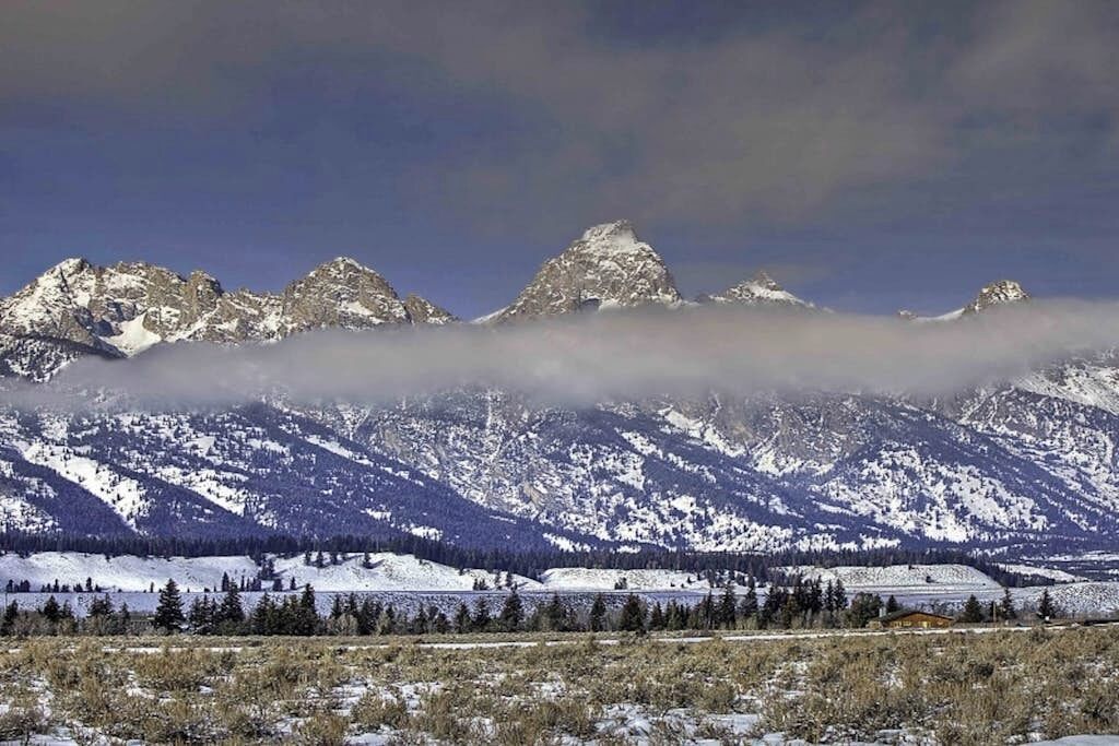 A mountainous landscape with snow-covered peaks, a forested valley, and a small building in the foreground.