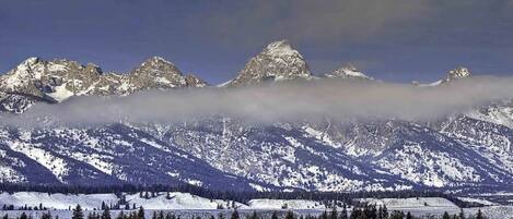 A mountainous landscape with snow-covered peaks, a forested valley, and a small building in the foreground.