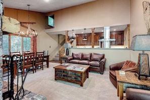 A spacious living area with a leather sofa, wooden coffee table, and a dining table. A kitchen with wooden cabinets and a chimney is visible in the background. - Beautiful condo with full kitchen, mountain views (Wilson)