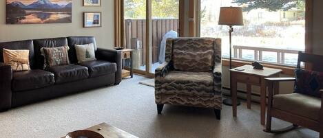 A living room with a sofa, armchair, coffee table, and a view through a sliding door.