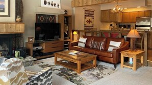 Ein Wohnzimmer mit Kamin, Fernseher, Sofa, Couchtisch und im Hintergrund eine Küchenzeile.