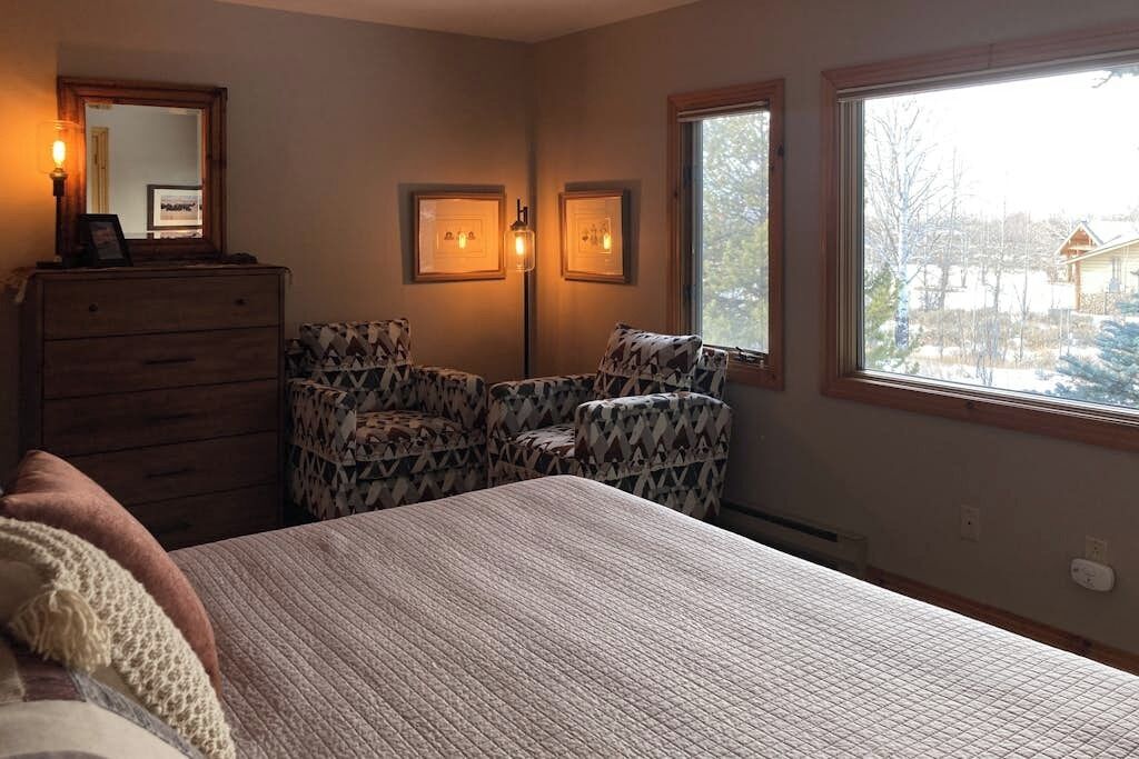 A bedroom with a bed, dresser, two chairs, two wall-mounted lights, and a window with a view.