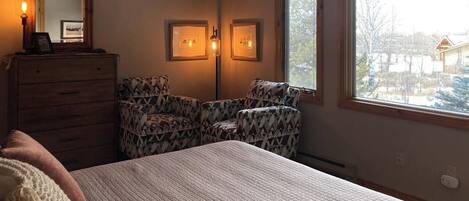 A bedroom with a bed, dresser, two chairs, two wall-mounted lights, and a window with a view.