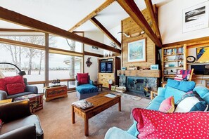 Un salon chaleureux avec une cheminée, des poutres apparentes, une bibliothèque et une grande fenêtre.
